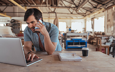 man in a workshop talking on a phone while looking at a laptop
